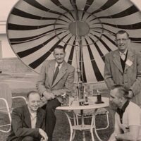 J. Leroy Smith standing at a table on the right of the table with glasses, William Penson on left sitting on the table, Howard McCully on left kneeling and Stan Plona on right in white shirt