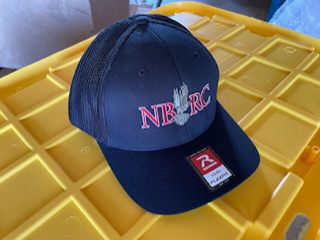 A black hat with the letters nbrc on it|A baseball cap with the words nrrc on it.|A hat sitting on top of yellow plastic trays.